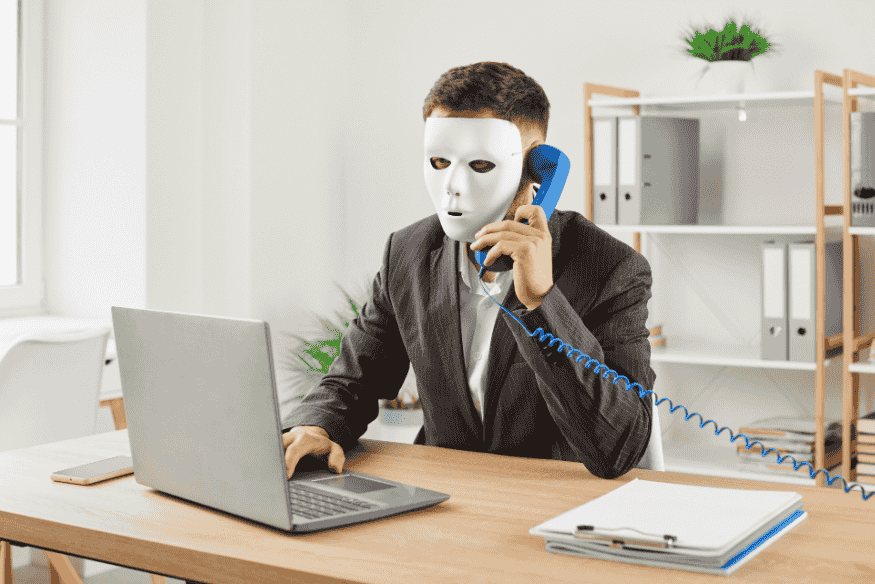 Homem usando máscara branca enquanto fala ao telefone e utiliza um notebook, simbolizando golpes e fraudes em negociações de dívidas.