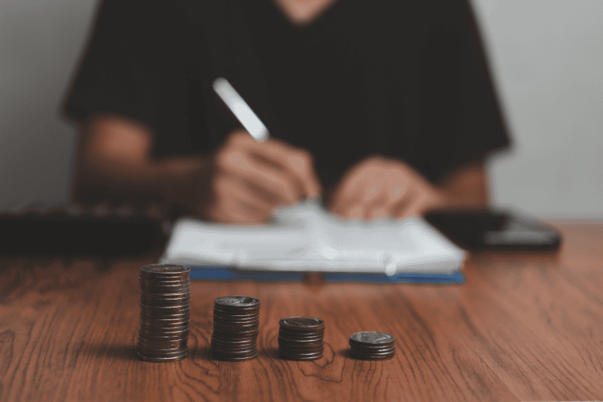 Pessoa fazendo anotações financeiras com pilhas de moedas organizadas em ordem crescente, representando o levantamento e organização das dívidas para planejar o pagamento.